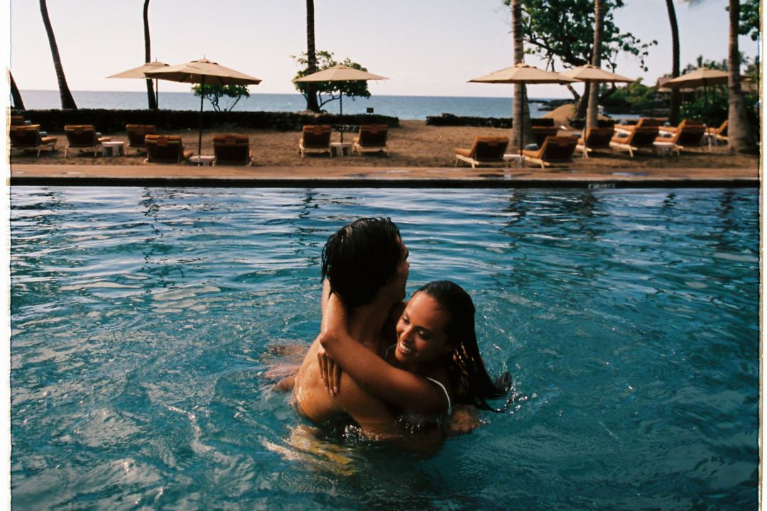 A couple embracing each other in a swimming pool with the beach and lounge chairs in the background.